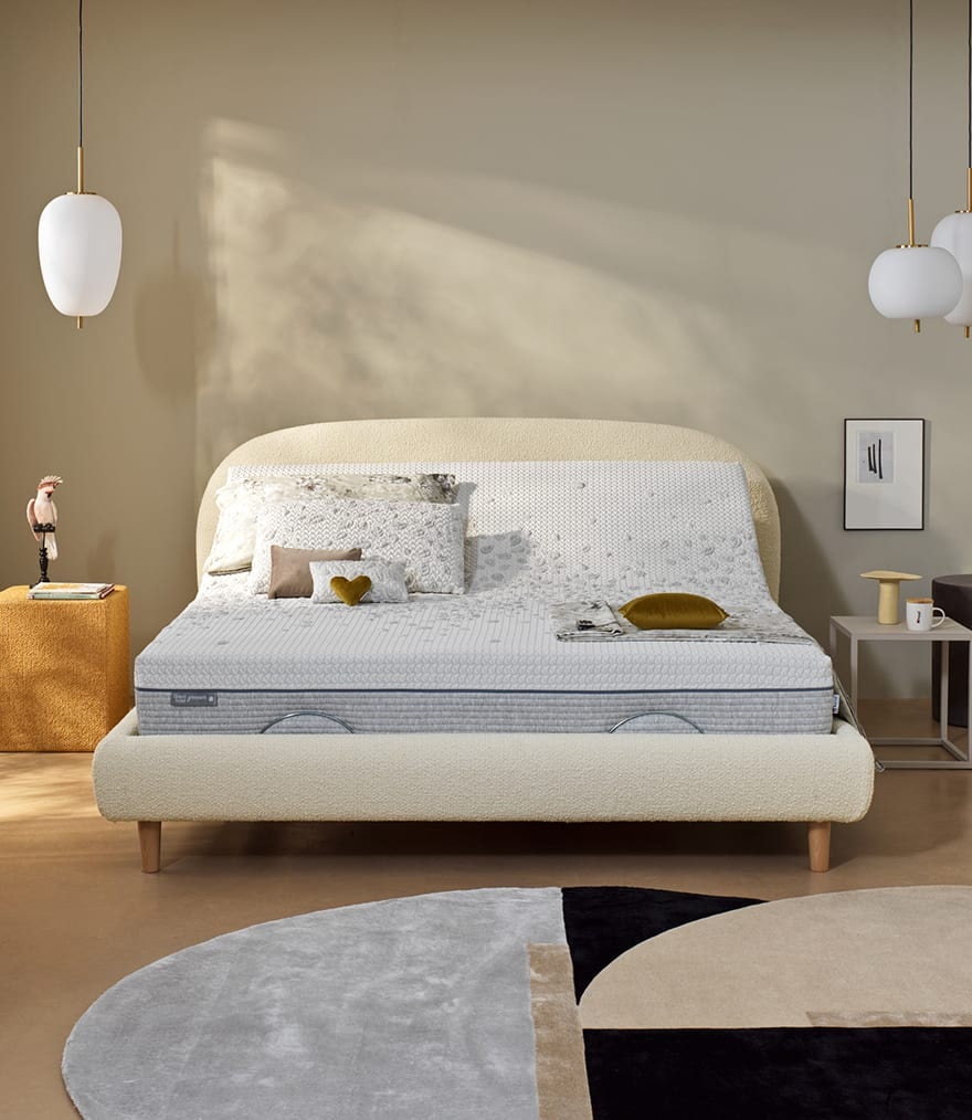 Letto con testiera e pediera imbottite color crema, cuscini decorativi, due comodini con lampade e tappeto geometrico toni…