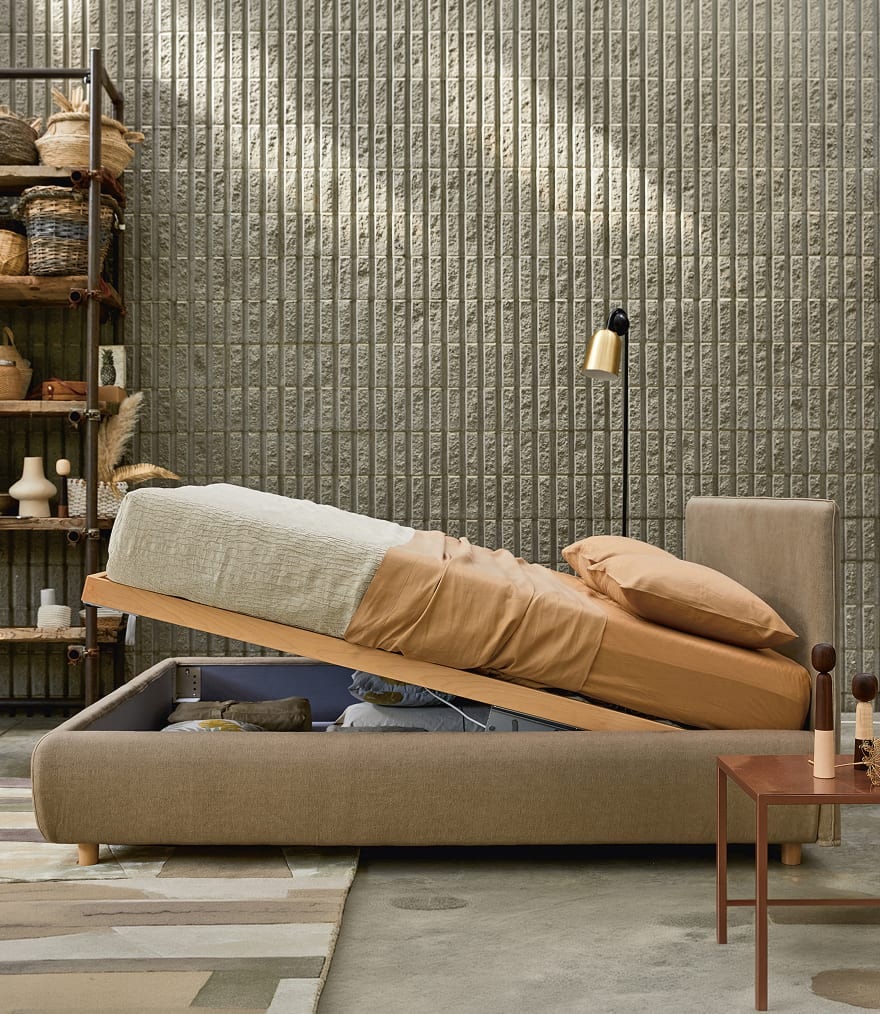 Letto contenitore beige con rete sollevata e biancheria arancione in stanza con parete in cemento e scaffale di vimini.