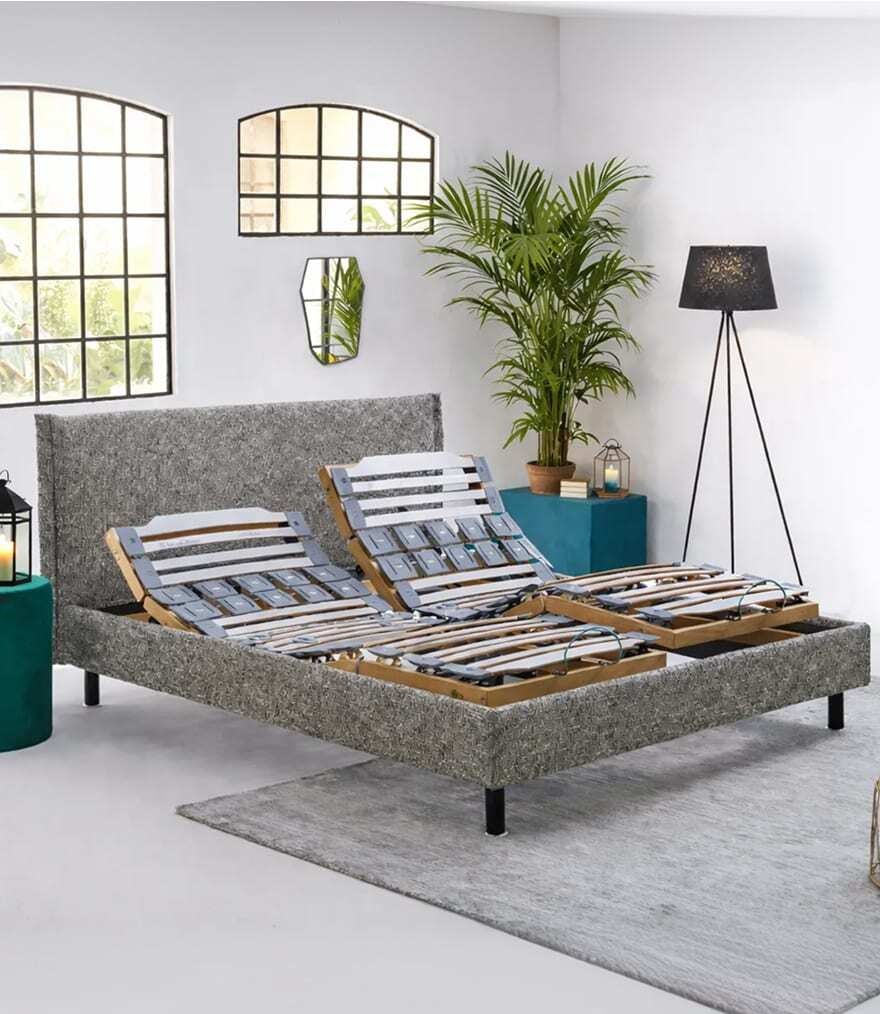 Letto matrimoniale con rete regolabile doppia in tessuto grigio, accanto pianta verde e lampada da terra nera.