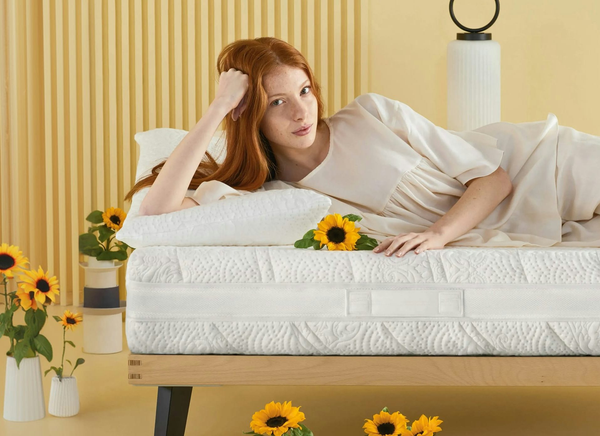 Donna dai capelli rossi stesa su un letto bianco con girasoli su un fondo giallo con vasi e lampada decorativa.