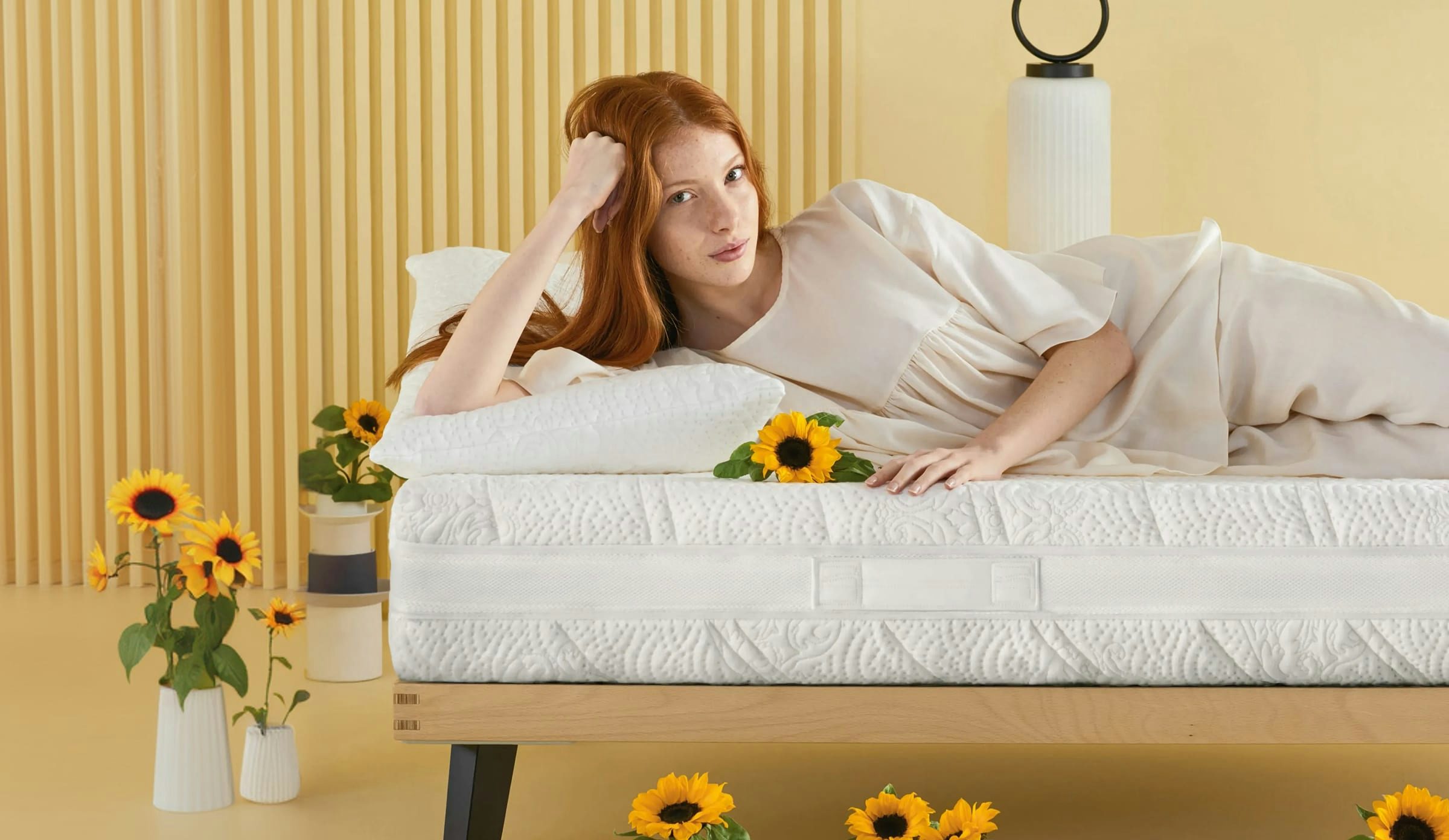 Donna dai capelli rossi stesa su un letto bianco con girasoli su un fondo giallo con vasi e lampada decorativa.