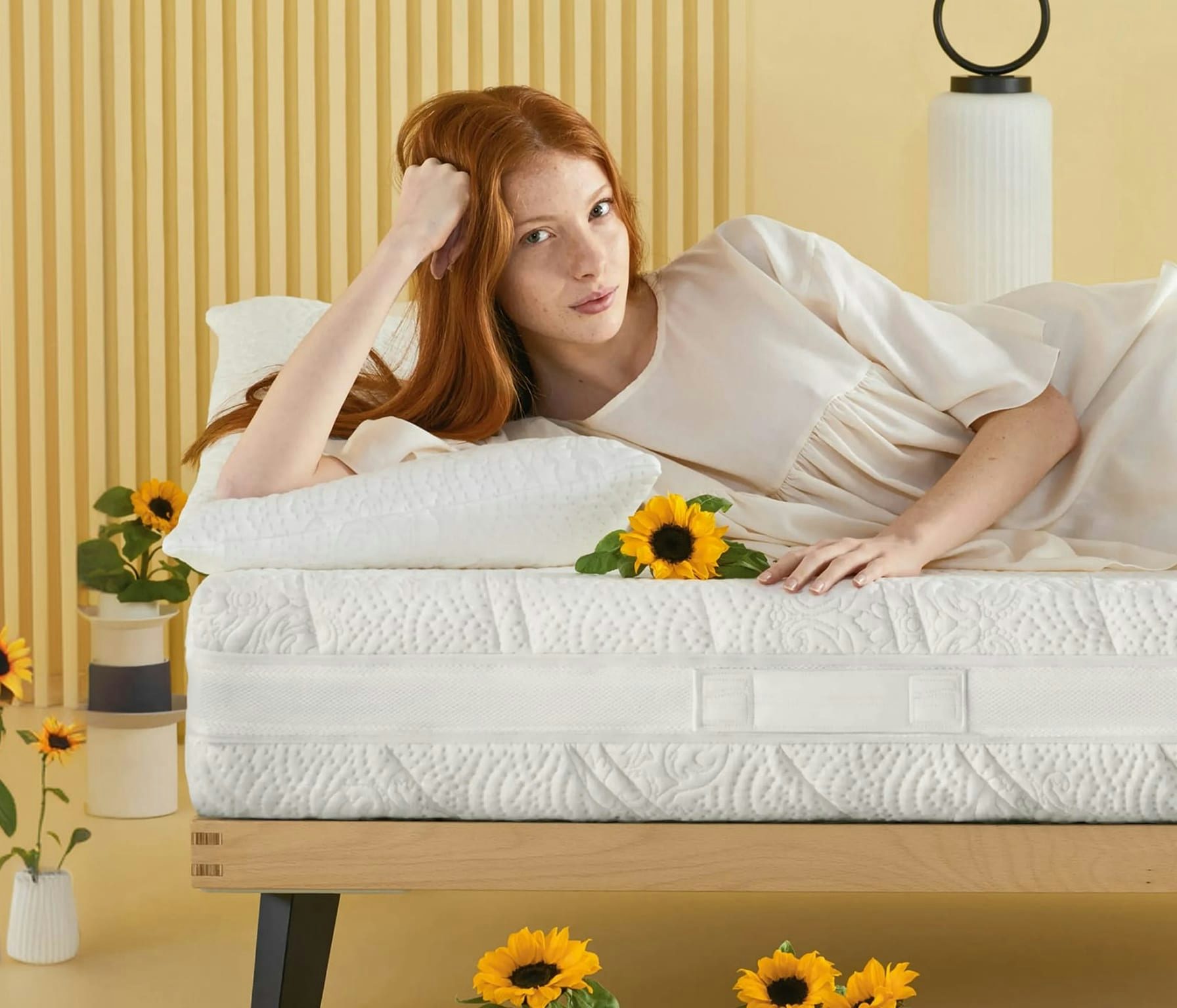 Donna con capelli rossi sdraiata su un letto con materasso bianco e fiori di girasole in una stanza dai toni chiari.