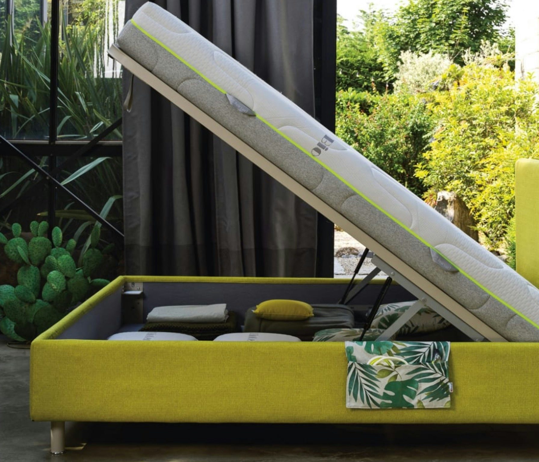 Letto contenitore giallo aperto con materasso sollevato, cuscini e coperte piegate all'interno, sfondo di piante verdi.