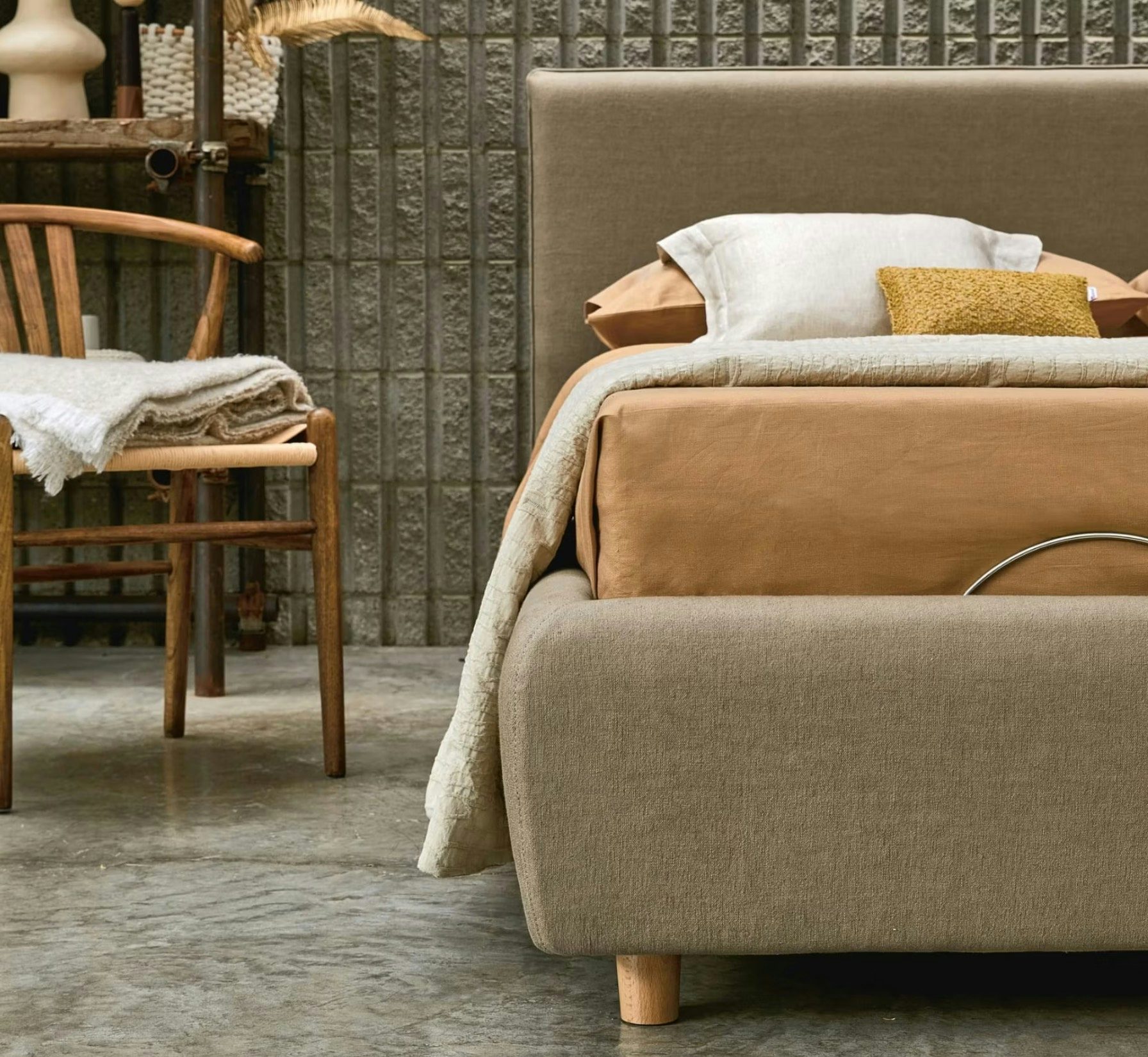 Letto beige imbottito con cuscini color ocra e bianco, sedia in legno con coperta chiara su pavimento cemento.
