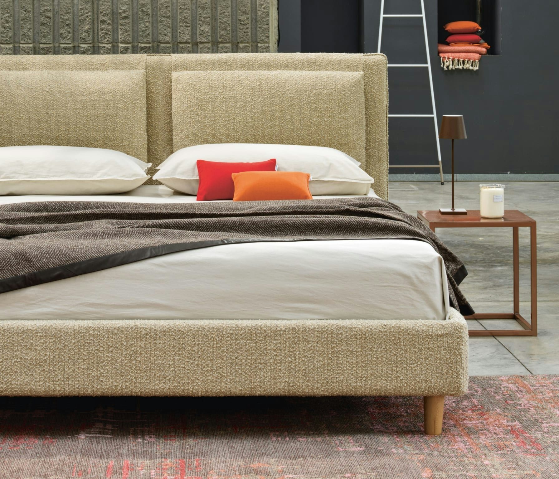 Letto imbottito beige con cuscini bianchi, due cuscini decorativi arancione e rosso, coperta marrone, lampada e candela sul…