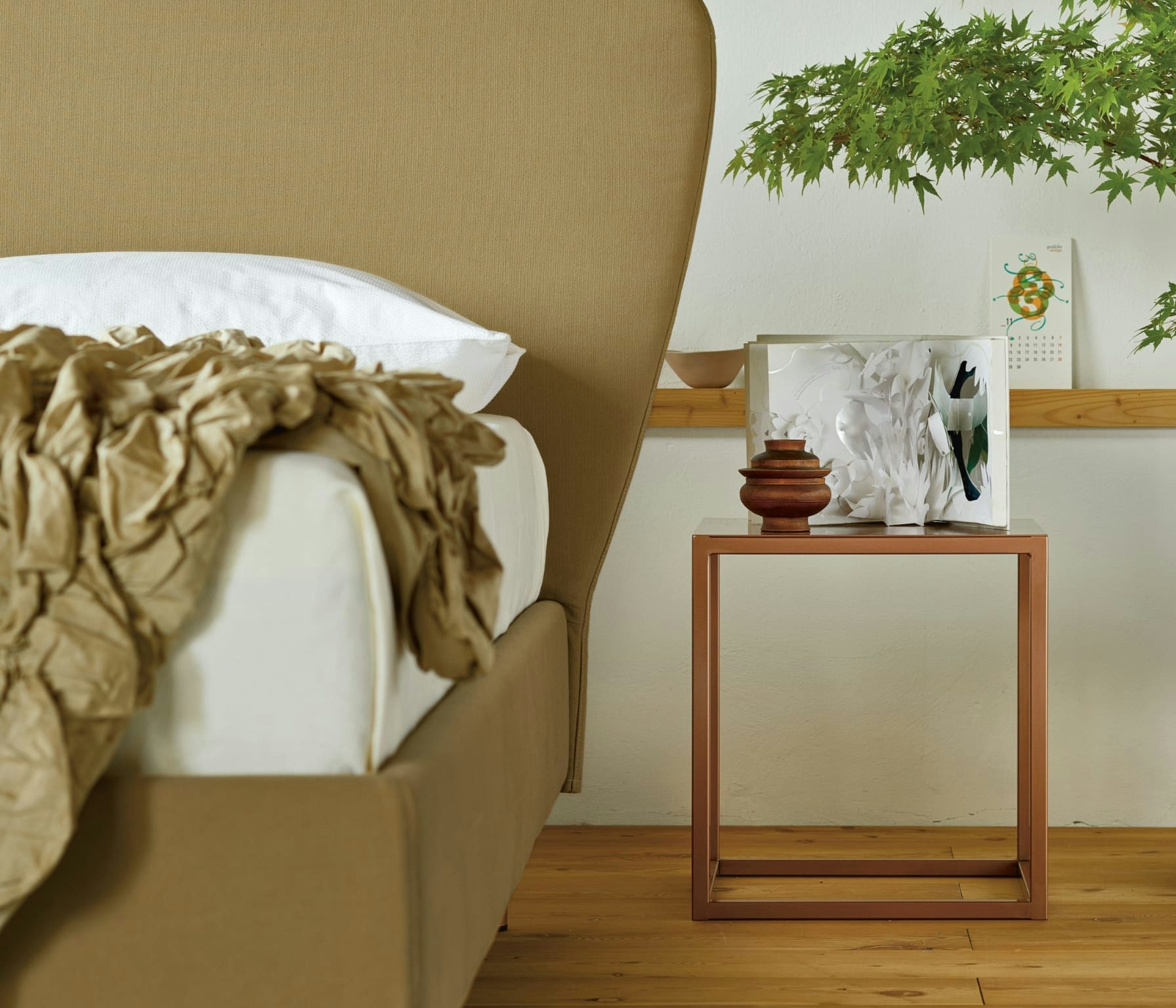 Letto beige con coperta stropicciata, comodino metallico con vaso in legno e libro aperto, parete bianca con pianta sopra.