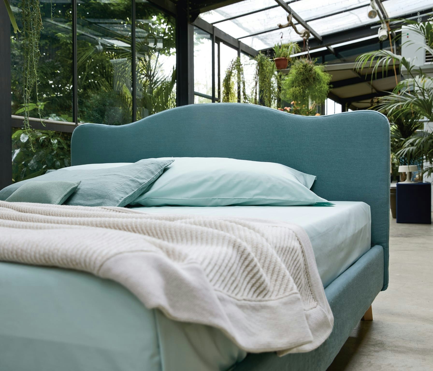 Letto con testata imbottita azzurra, cuscini e coperta beige in ambiente luminoso con molte piante verdi.