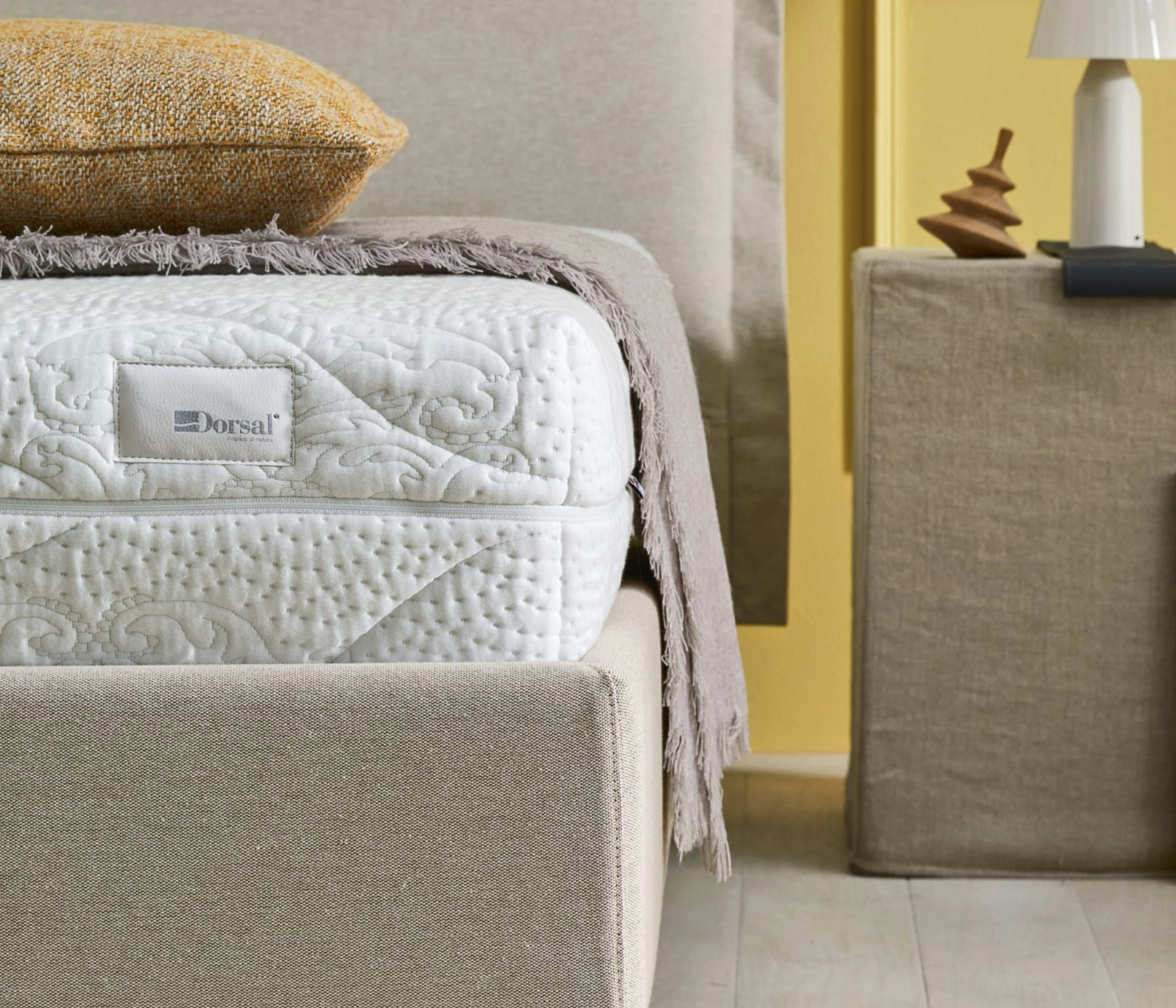 Letto imbottito beige con cuscino giallo, materasso bianco Dorsal e copriletto grigio chiaro accanto a comodino beige.