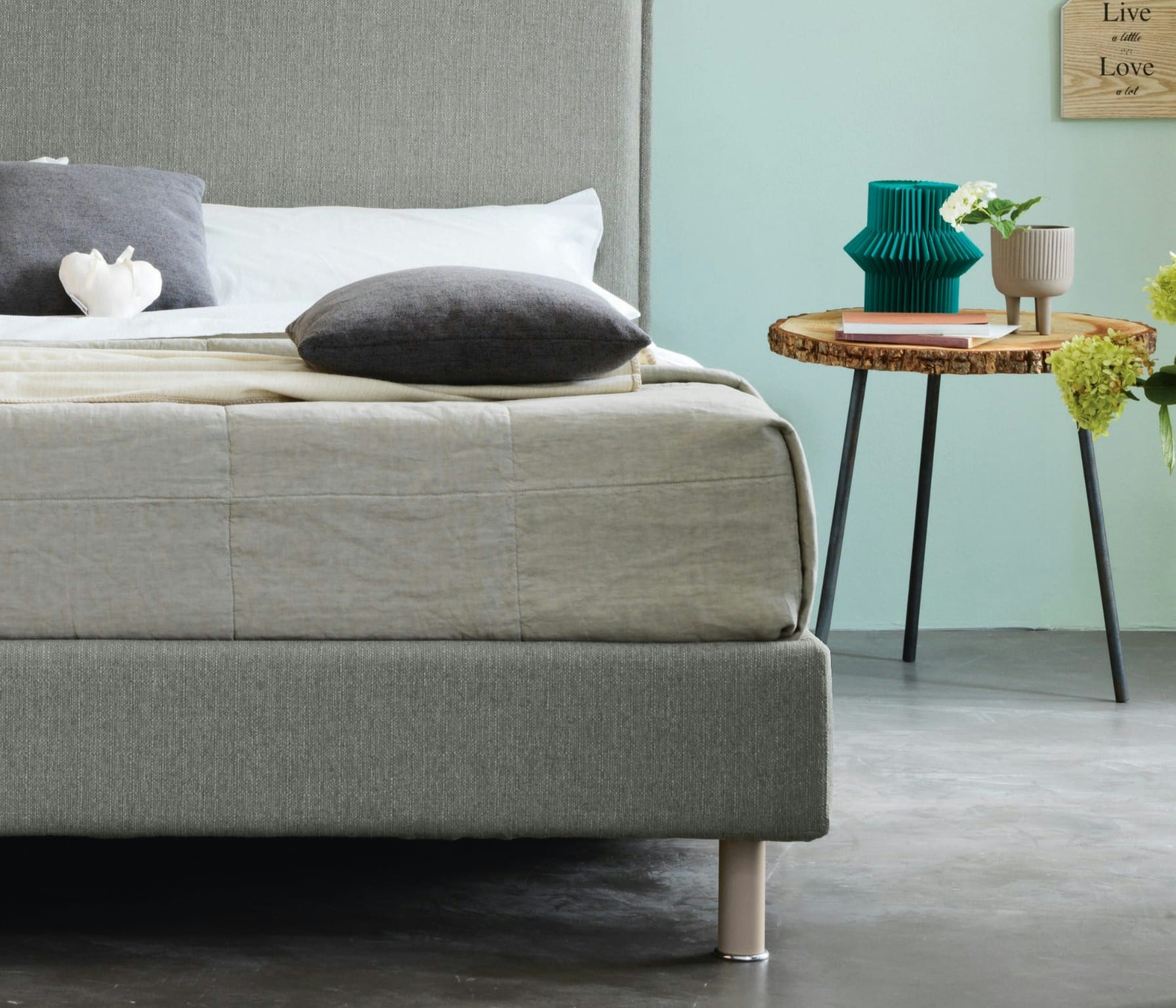 Letto matrimoniale con testiera e struttura grigia, cuscini grigi e bianchi, tavolino in legno con vaso e libri su pavimento…