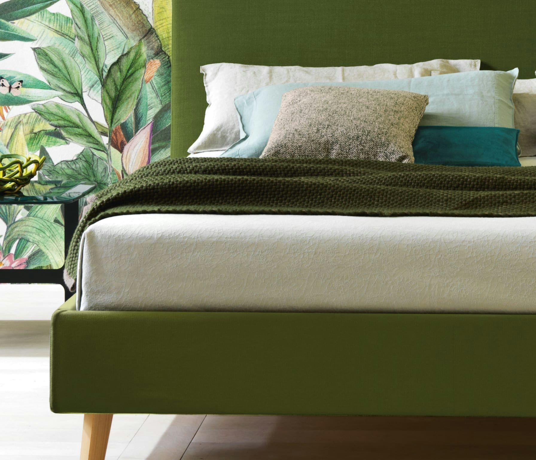 Letto con struttura verde, coperta verde scuro, cuscini beige, azzurri e blu, sfondo con decorazione foglie verdi e farfalle…