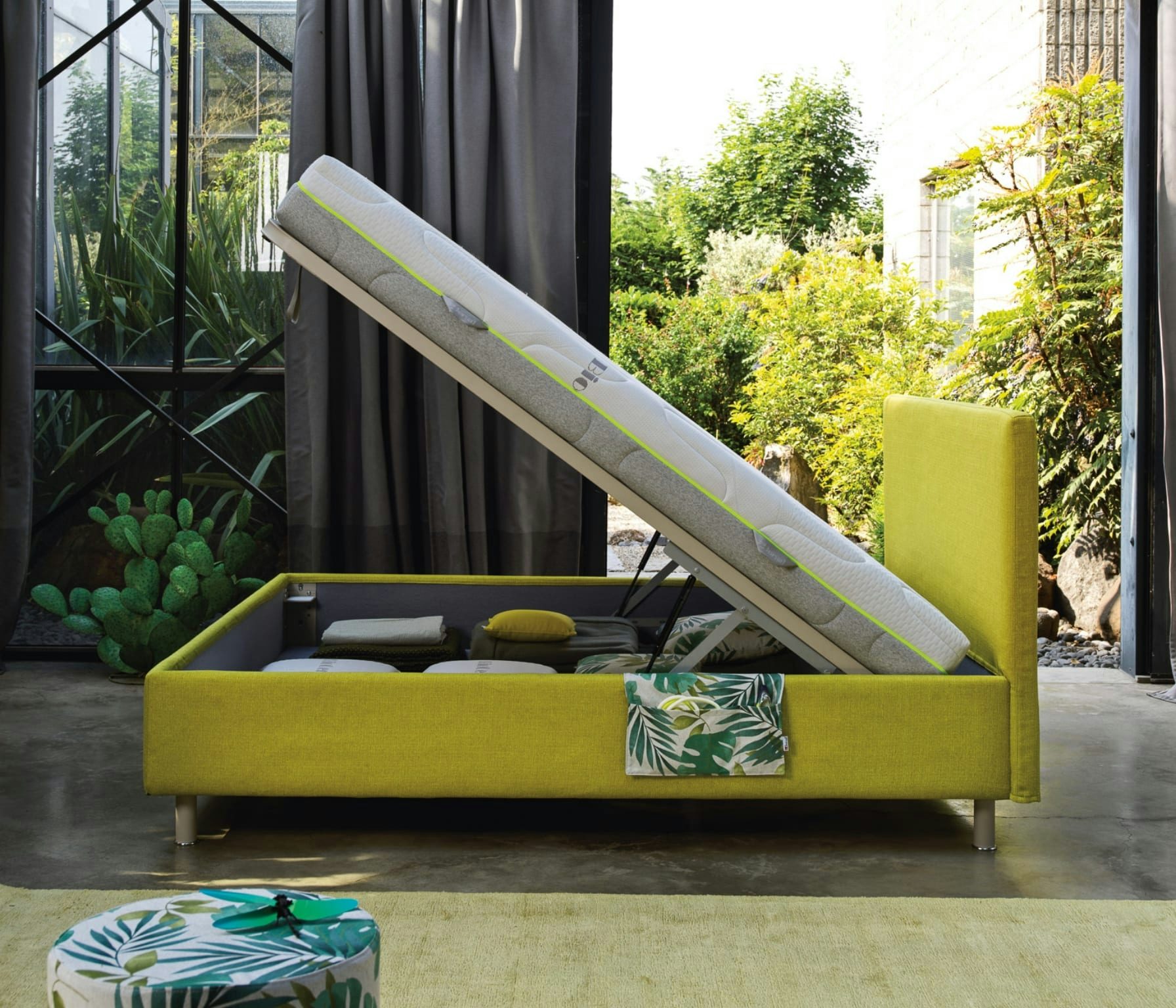 Letto contenitore verde aperto con materasso sollevato, cuscini e coperte all'interno, stanza con grandi finestre e vista su…