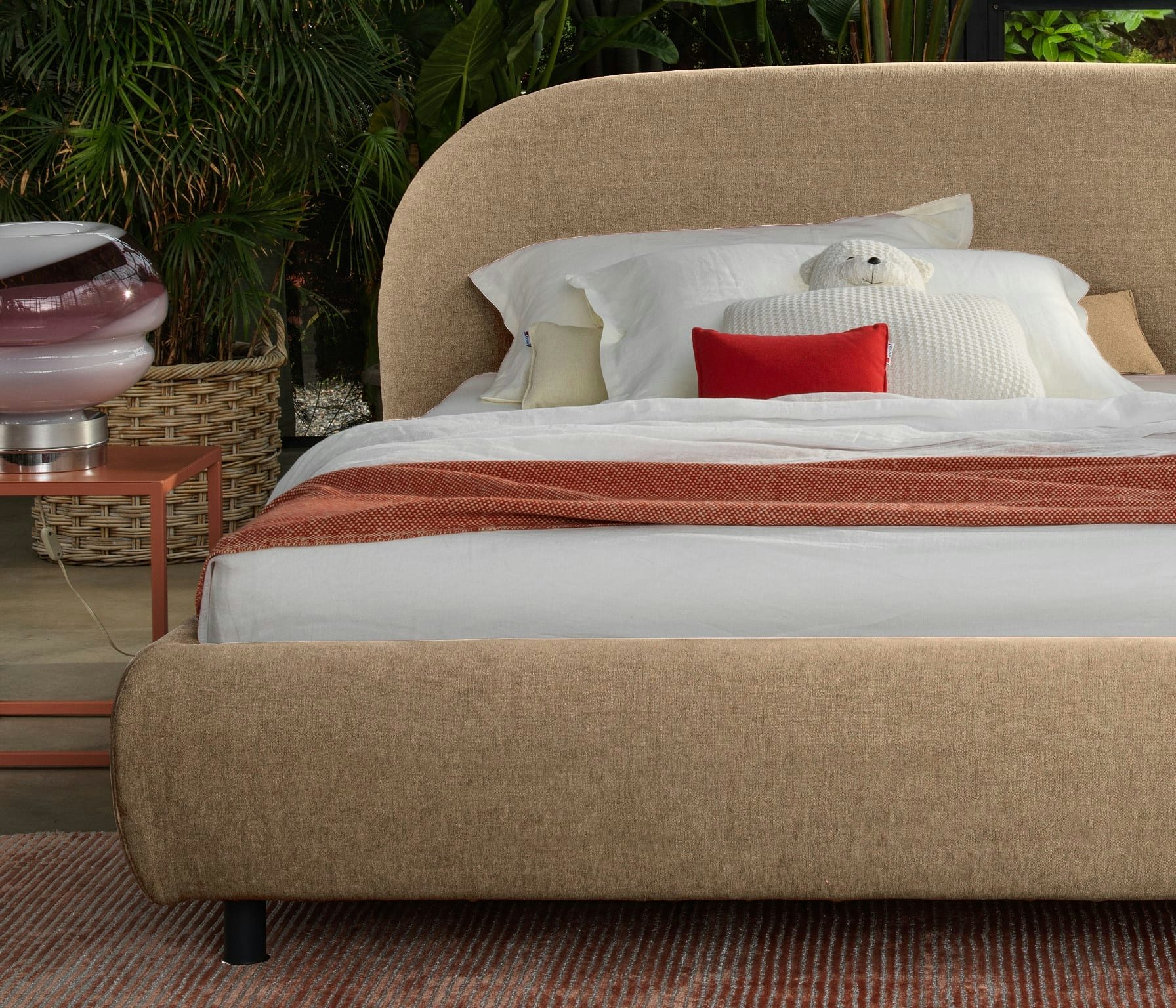 Letto beige imbottito con cuscini bianchi, un cuscino rosso e un peluche orso, accanto pianta in vaso e lampada rosa.