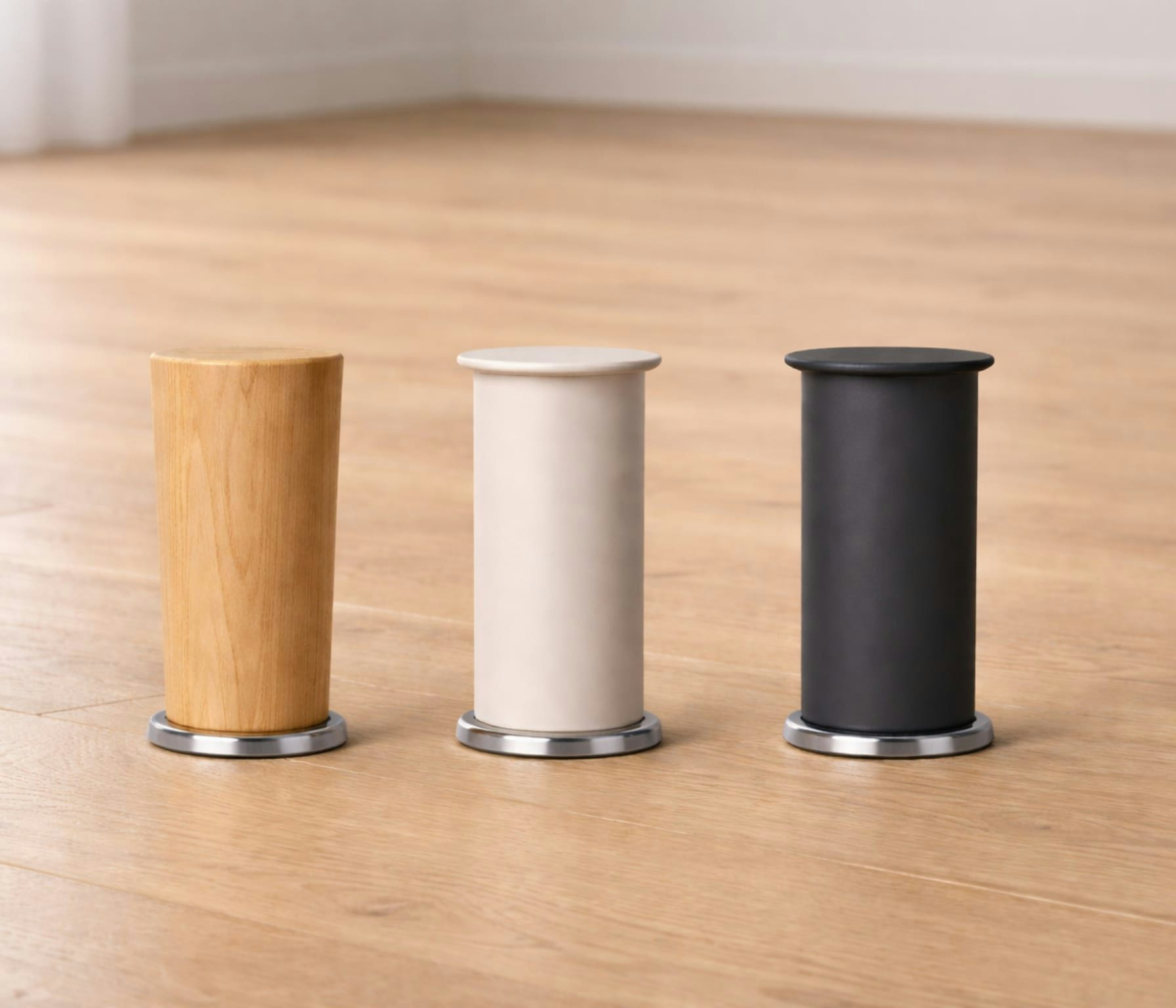 Tre gambe di diversi colori per mobili, disposte su un pavimento in legno chiaro, con base metallica tonda e parte superiore…