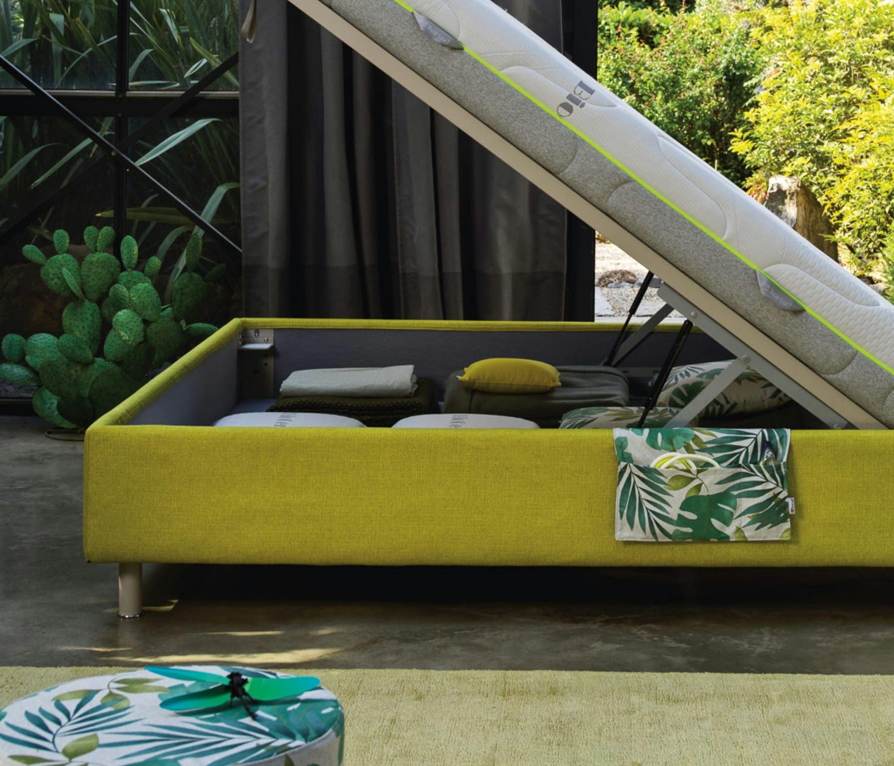 Letto contenitore verde aperto con cuscini, plaid e coperte, con pouf coordinato in primo piano e piante sullo sfondo.