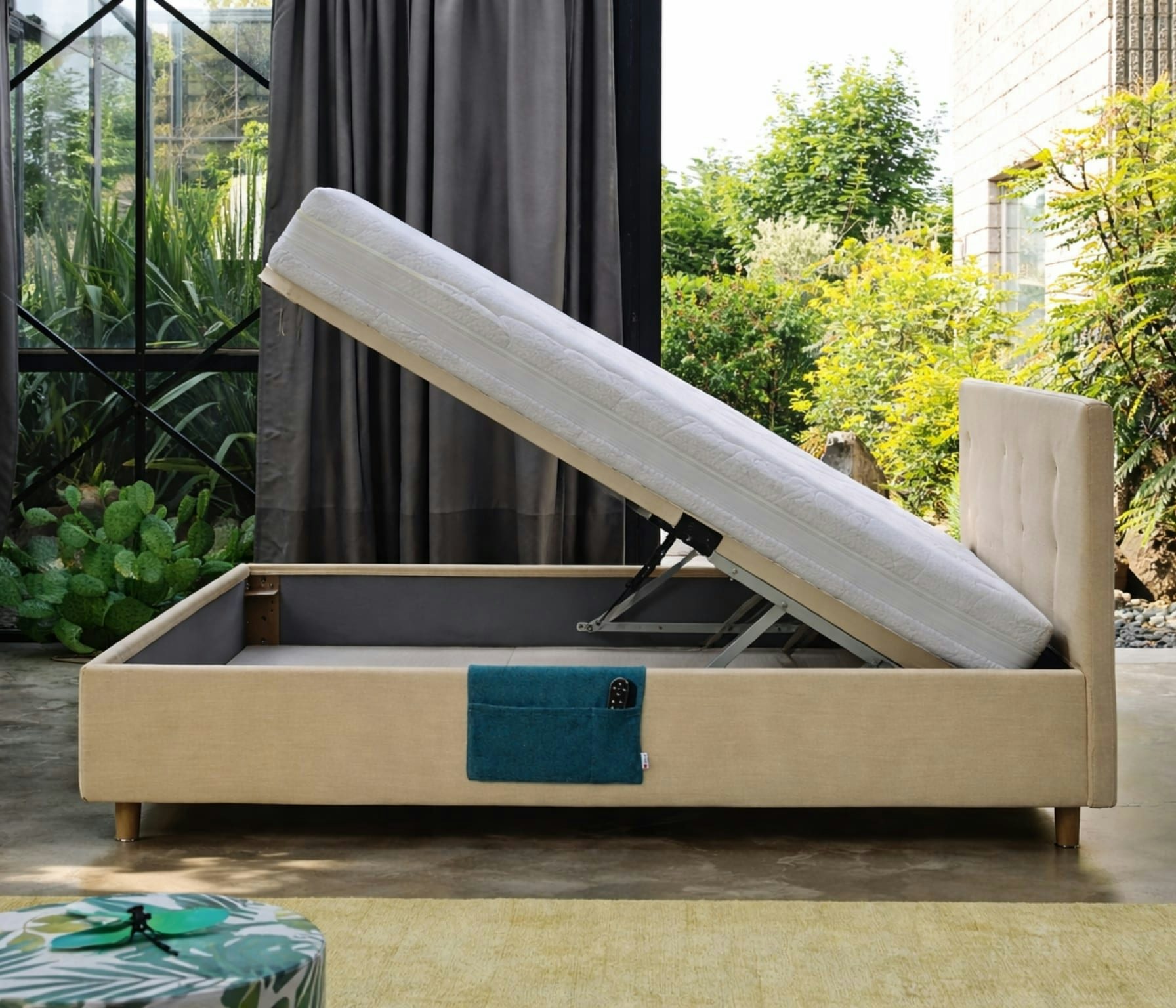 Letto contenitore beige con rete sollevabile e materasso bianco, tasca blu con telecomando, pavimento in cemento e vetrata…