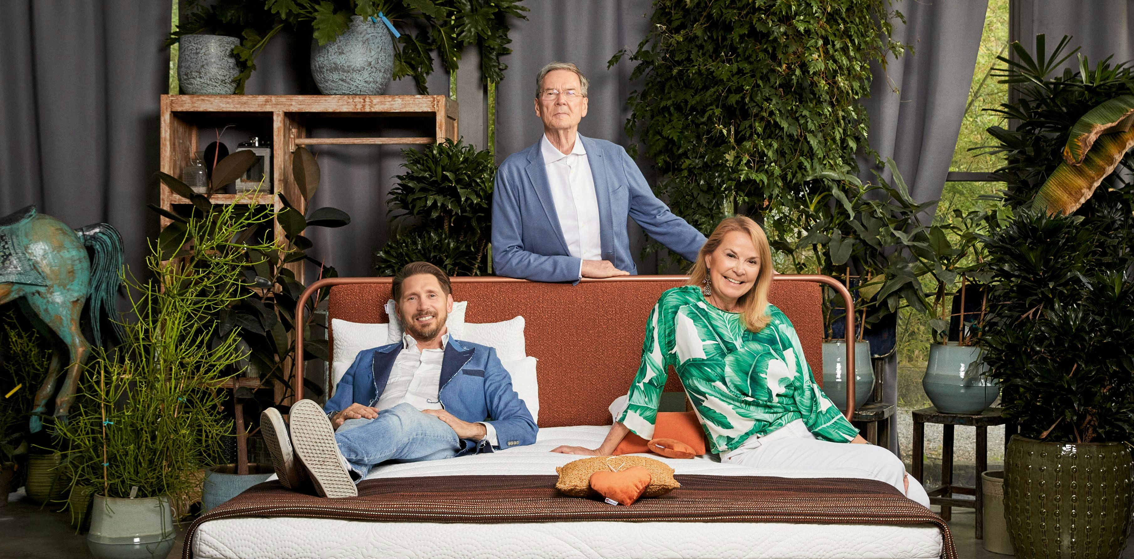 Tre persone posano sorridenti su un letto matrimoniale in una stanza con molte piante verdi e decorazioni rustiche.