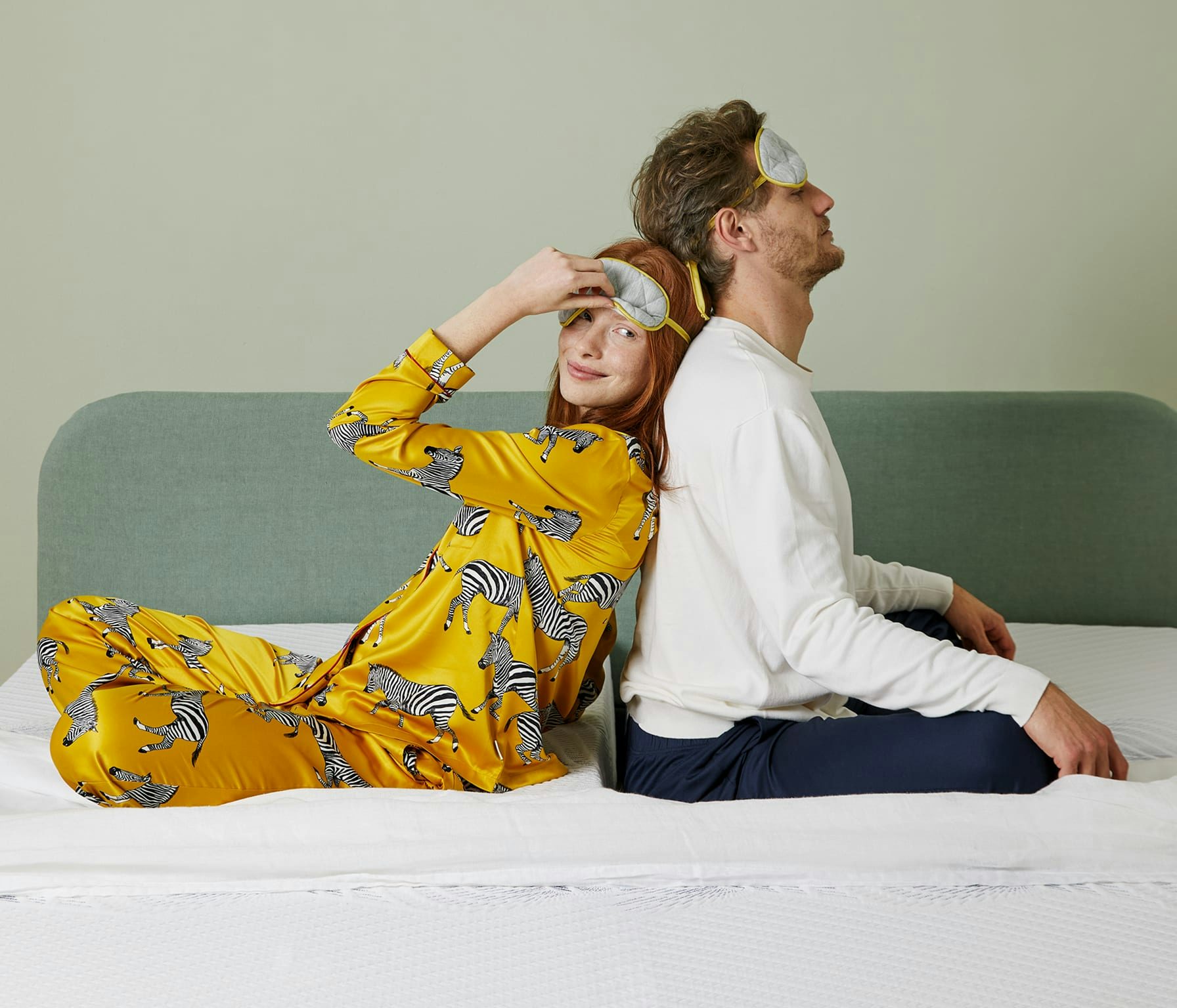 Coppia seduta sul letto schiena contro schiena, lei con pigiama giallo a zebre e mascherina da notte, lui con maglia bianca…
