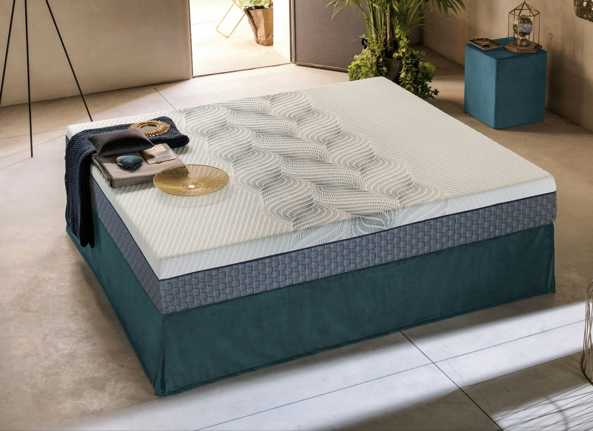 Letto con base imbottita blu petrolio, materasso bianco con motivi geometrici grigi, coperta e oggetti decorativi sopra.