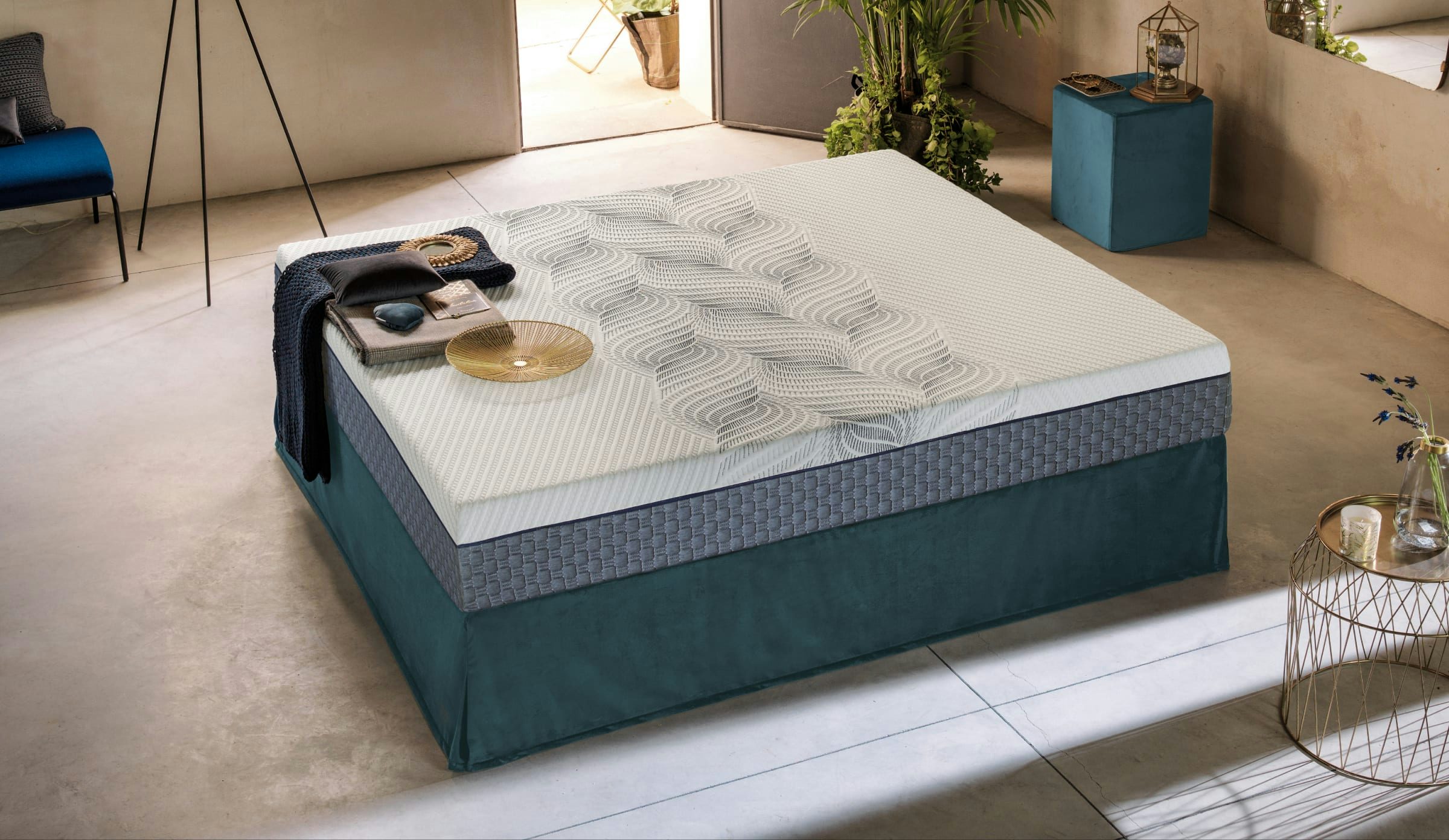 Letto con base imbottita blu petrolio, materasso bianco con motivi geometrici grigi, coperta e oggetti decorativi sopra.