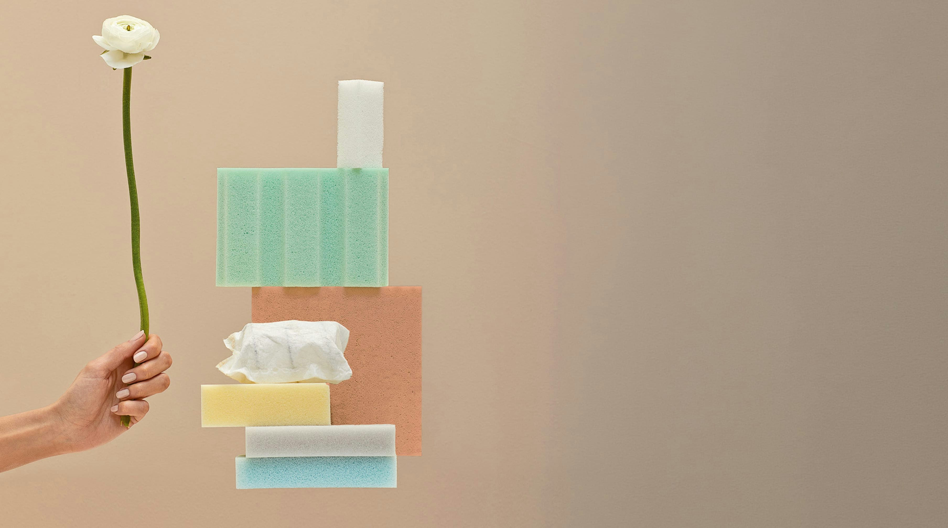 Mano con unghie curate che tiene un fiore bianco sopra una composizione di spugne colorate su sfondo beige.