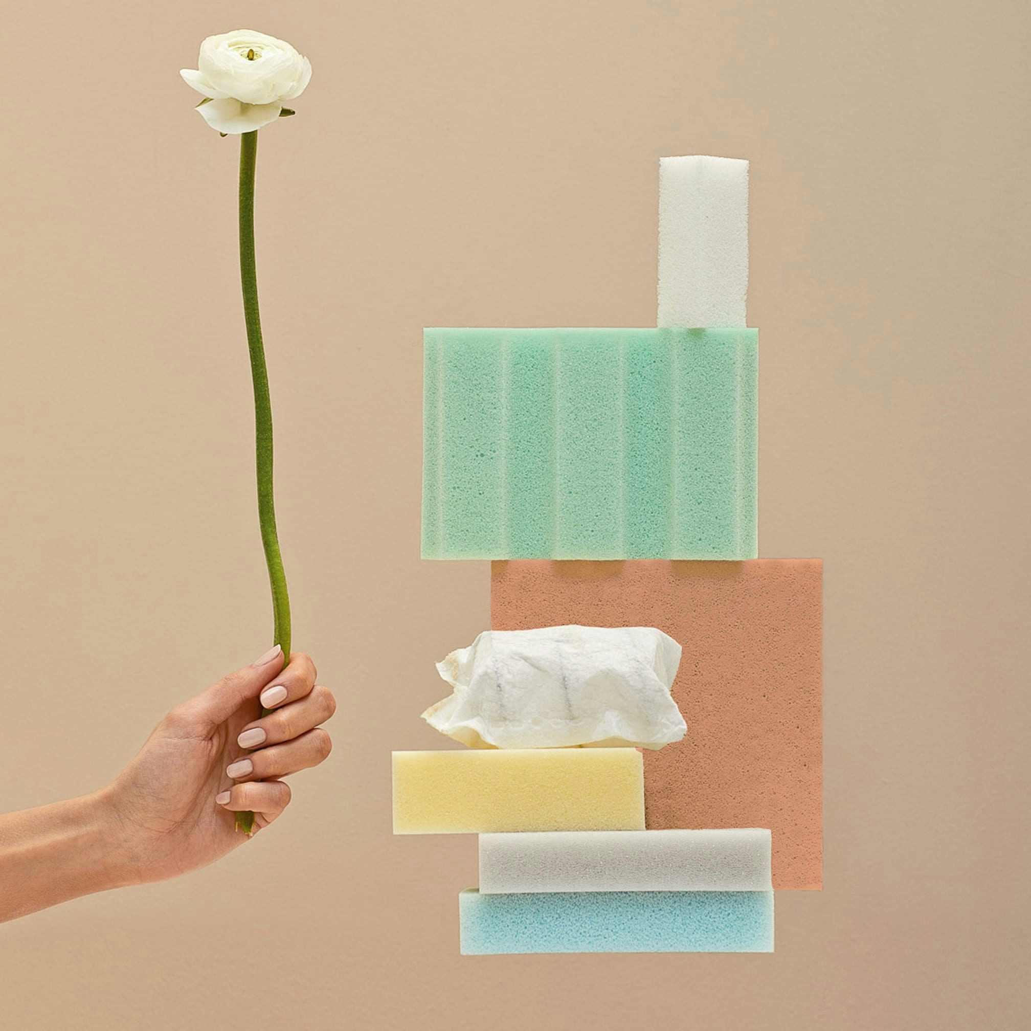 Mano con unghie curate che tiene un fiore bianco sopra una composizione di spugne colorate su sfondo beige.