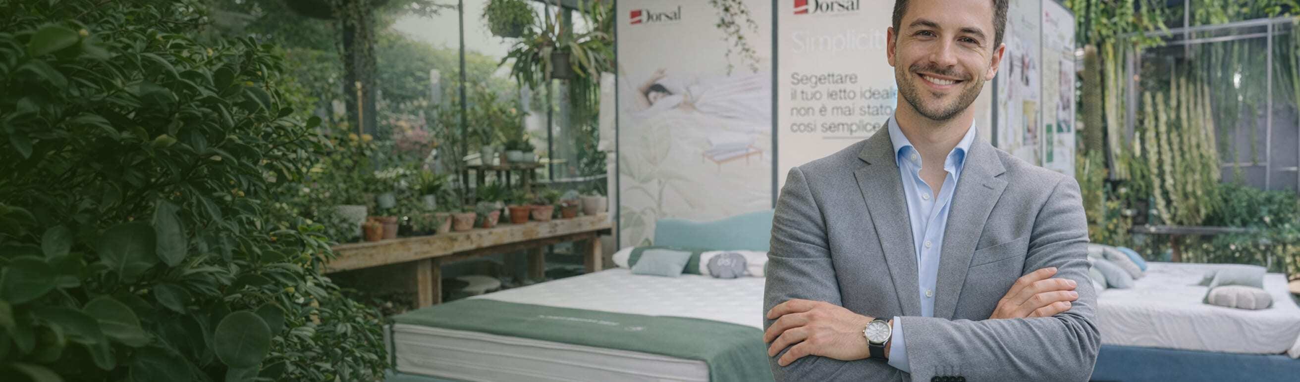 Uomo sorridente in giacca grigia con braccia incrociate, circondato da piante e letti in esposizione.