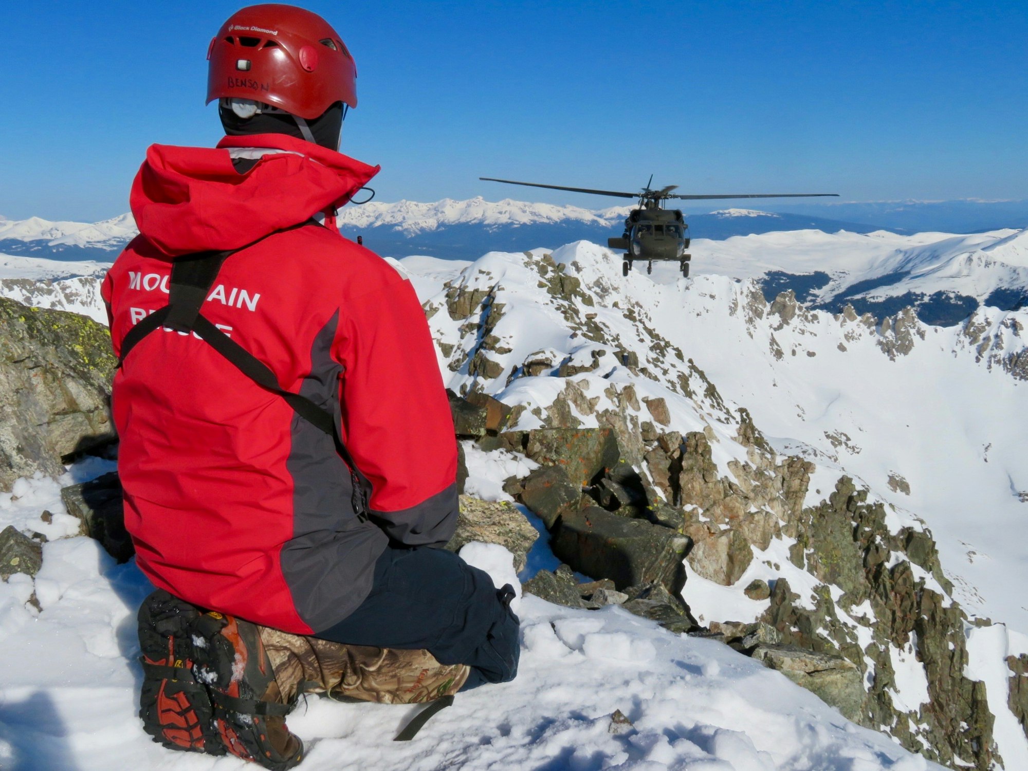 Search and Rescue Volunteer kneels on a high mountain ridge looking out at a helicopter