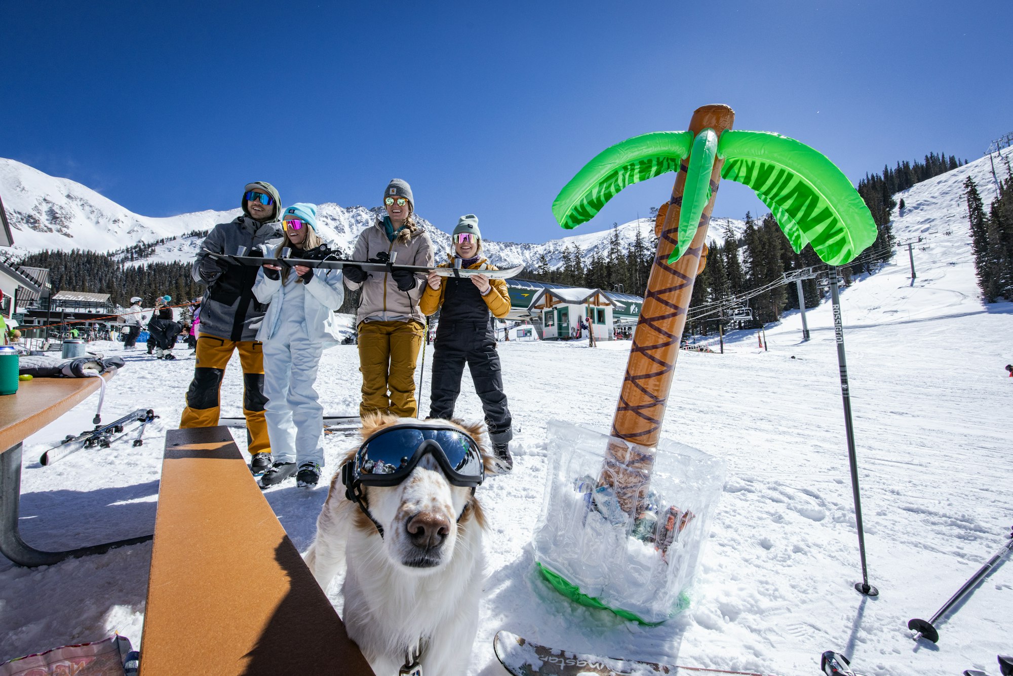 A dog and four skiers pose on the snow at the base of Arapahoe Basin