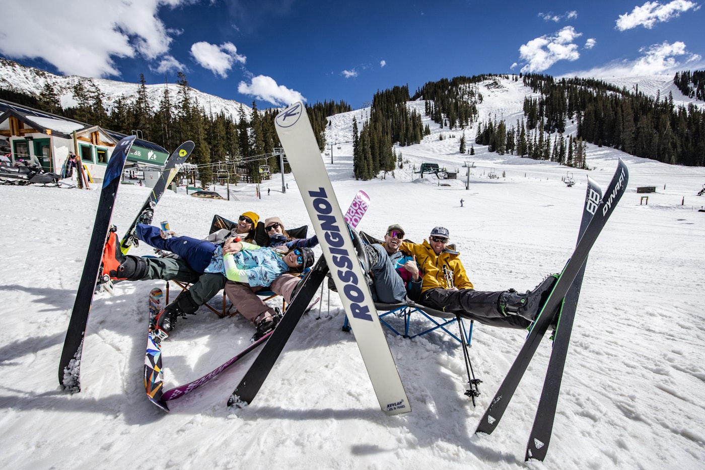 Five skiers sit in fold out chairs on the snow at the base of Arapahoe Basin