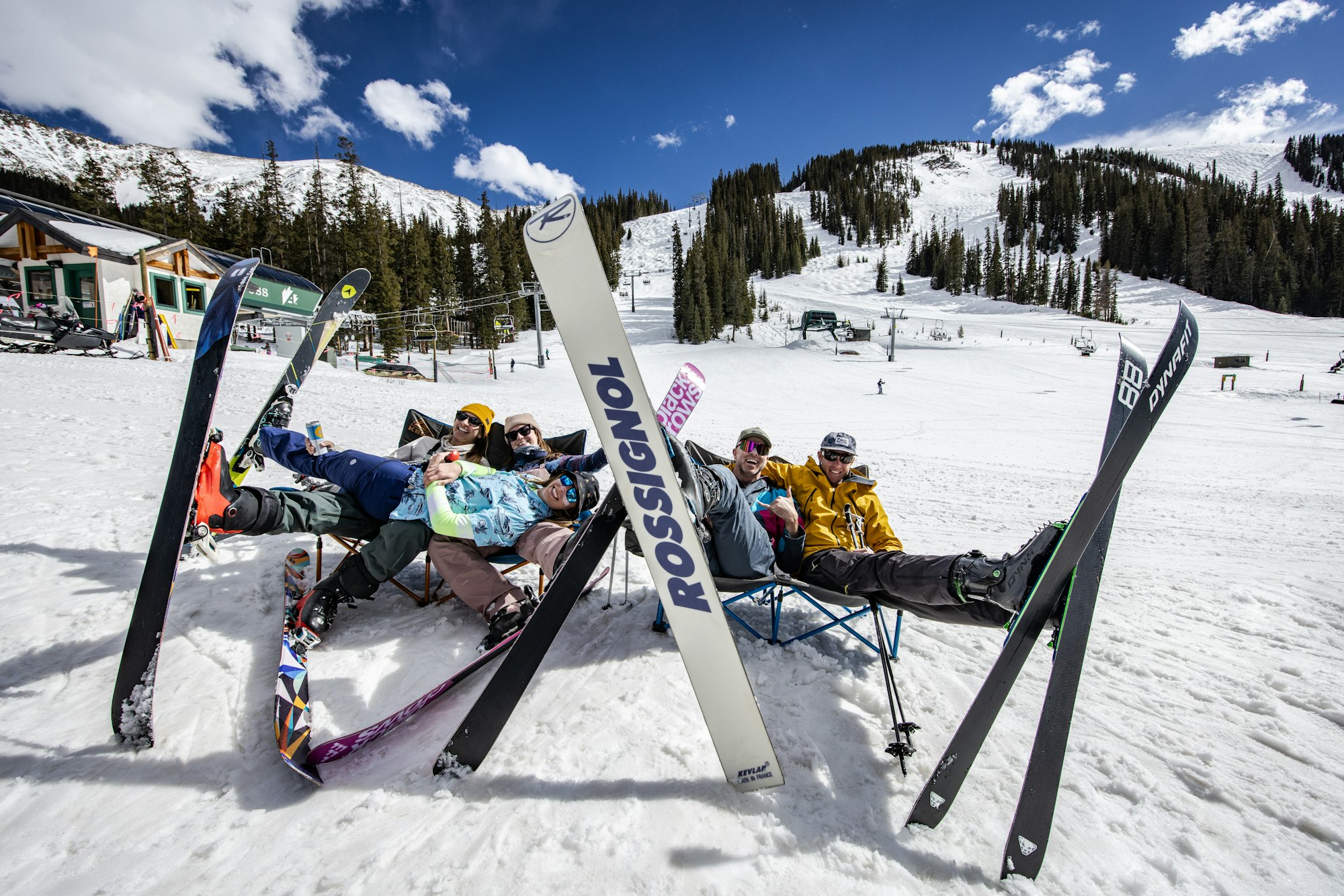Five skiers sit in fold out chairs on the snow at the base of Arapahoe Basin