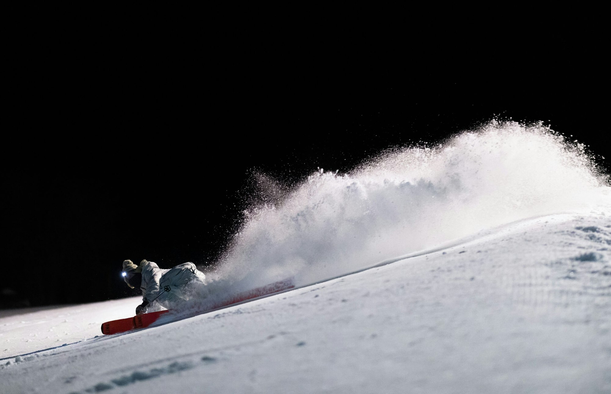 Skier carves down a groomed trail at night spraying snow behind them