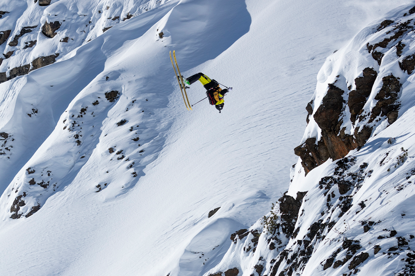 Skier does a backflip off of a rocky and snowy mountain face