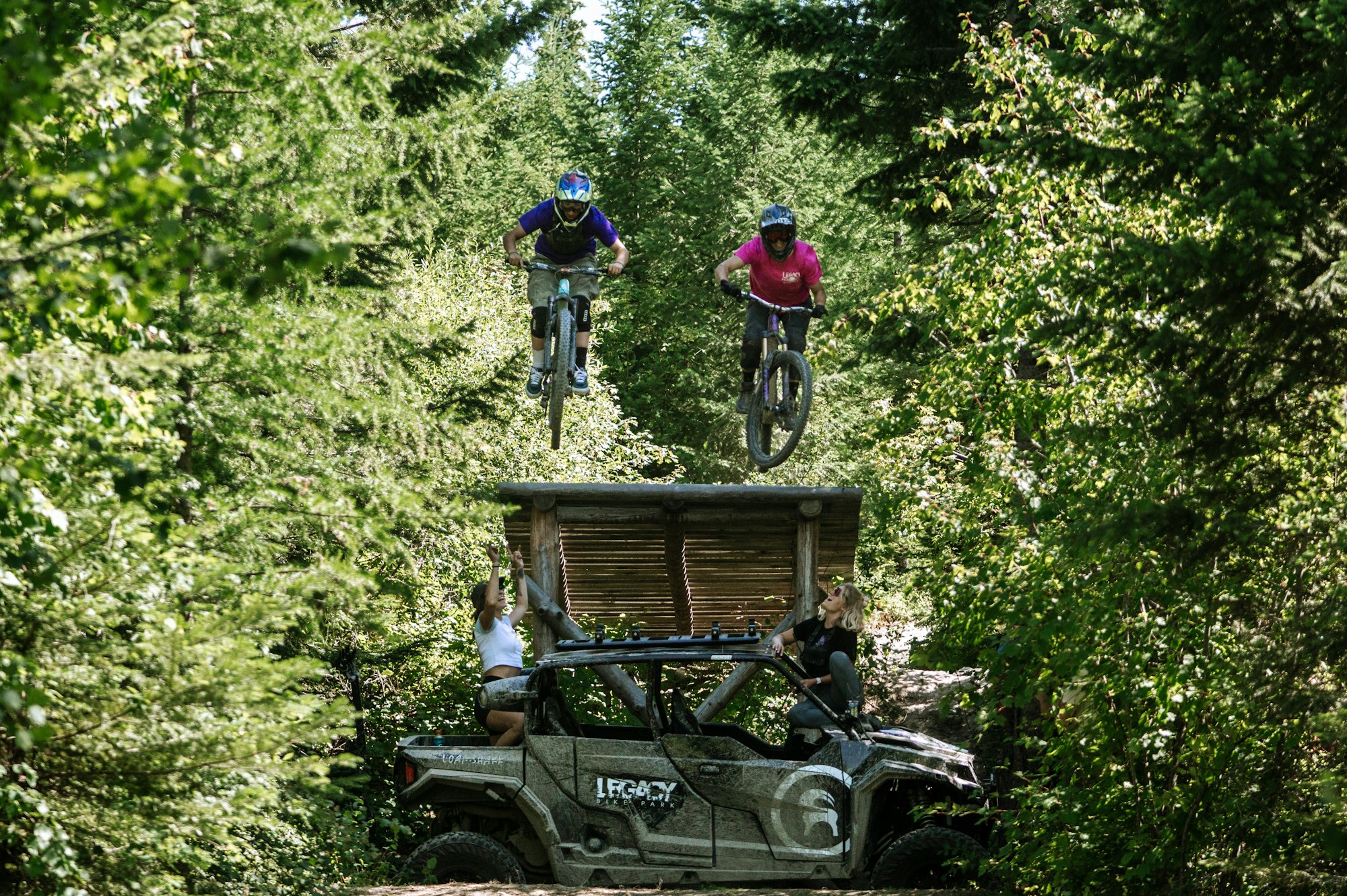 Women being cheered on as they hit a jump together at Legacy Bike Park .