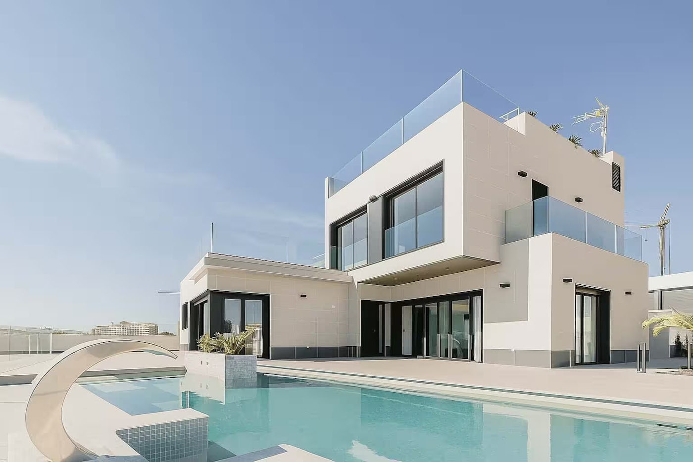 Modern two-story luxury home with large glass windows, balconies, and a pool under a clear blue sky.