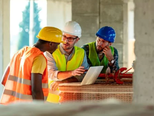 3 construction workers stood around a laptop in a construction site. They are all wearing PPE.