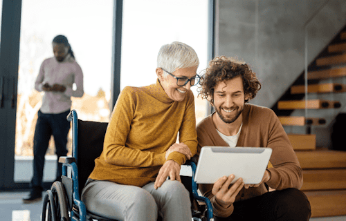 A silver-haired woman in a wheelchair looking at something on a tablet with a colleague.