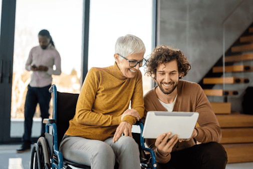 A silver-haired woman in a wheelchair looking at something on a tablet with a colleague.