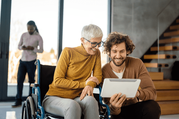 A silver-haired woman in a wheelchair looking at something on a tablet with a colleague.