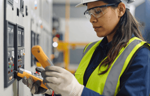 A female technician wearing safety gear inspects electrical control panels using testing devices in an industrial setting.
