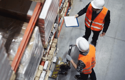 Two employees in hard hats and safety vests discussing safe lifting procedures in a warehouse.