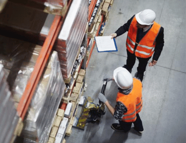 Two employees in hard hats and safety vests discussing safe lifting procedures in a warehouse.