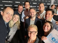 A group of eight people pose and smile for a photo at an indoor event, with a branded backdrop and certificate visible.