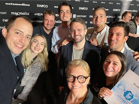 A group of eight people pose and smile for a photo at an indoor event, with a branded backdrop and certificate visible.