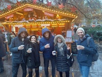 Five people in winter coats stand smiling and holding drinks in front of a festive outdoor market stall decorated with lights and holiday ornaments.