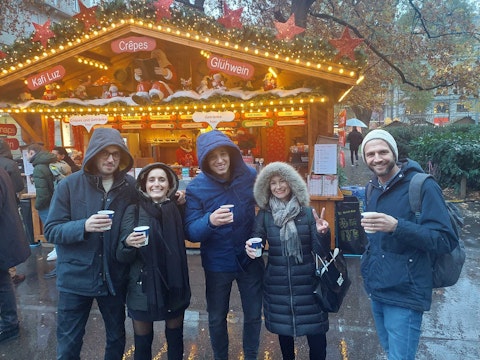 Five people in winter coats stand smiling and holding drinks in front of a festive outdoor market stall decorated with lights and holiday ornaments.