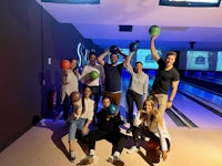 Eight people pose at a bowling alley, each holding a bowling ball up, with bowling lanes and score screens visible in the background.