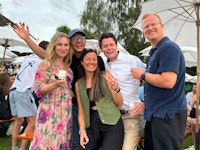 Five people standing close together outdoors at an event, smiling at the camera, with white umbrellas and other attendees in the background.