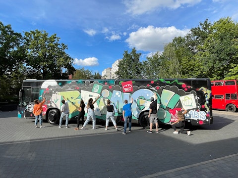 A group of people stands in front of a large black bus covered in colorful graffiti, with trees and a red double-decker bus in the background.