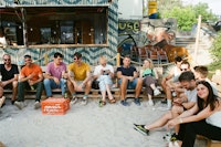 A group of people sit on benches in a sandy outdoor area, some holding drinks, in front of a bar with graffiti and stacked chairs in the background.