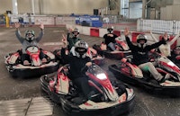 A group of five people in helmets and masks sit in red and white go-karts indoors, raising their hands and posing before a race.